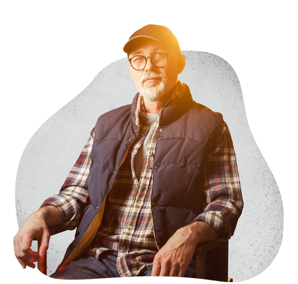 The Greenhouse Arts & Media Inc. - cropped image of man in directors chair, shown from waist up in flannel shirt, navy vest, with hat and glasses